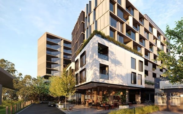 Modern multi story residential building built by GRC Buxton with a mix of white and natural-toned façade materials, featuring balconies and large windows. The building sits on a tree-lined corner with landscaped greenery and a street-level café with outdoor seating.