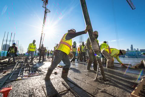 Power Construction workers pouring concrete over rebar on a jobsite with PPE and boots and other PPE on.