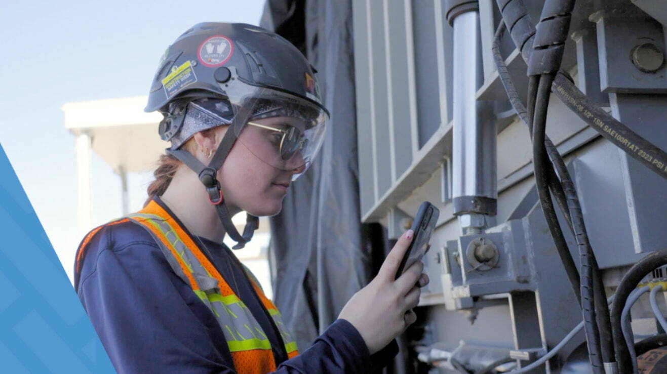 Construction worker taking a picture with phone wearing PPT and standing near heavy equipment.