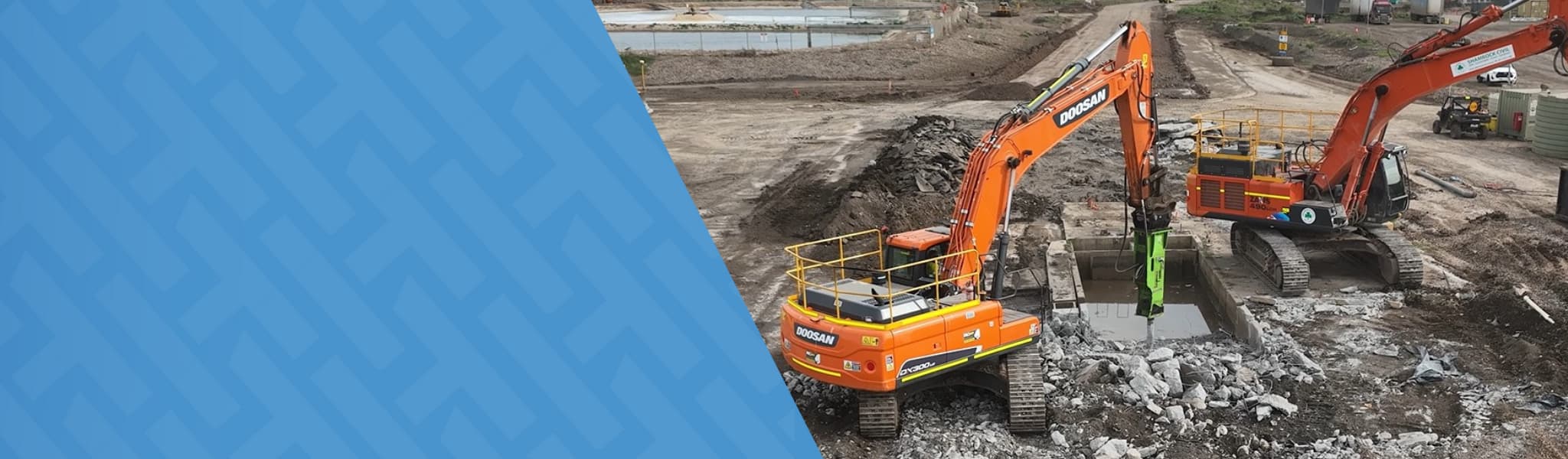 An orange excavator operates on a construction site, moving debris while a worker in high visibility clothing stands nearby overseeing the excavation work.