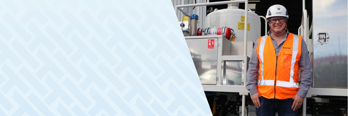 Man in high-visibility orange vest and hard hat standing beside a road marking machine, smiling on a construction site. There is light blue branded on the left with the logo of construction safety management software HammerTech.